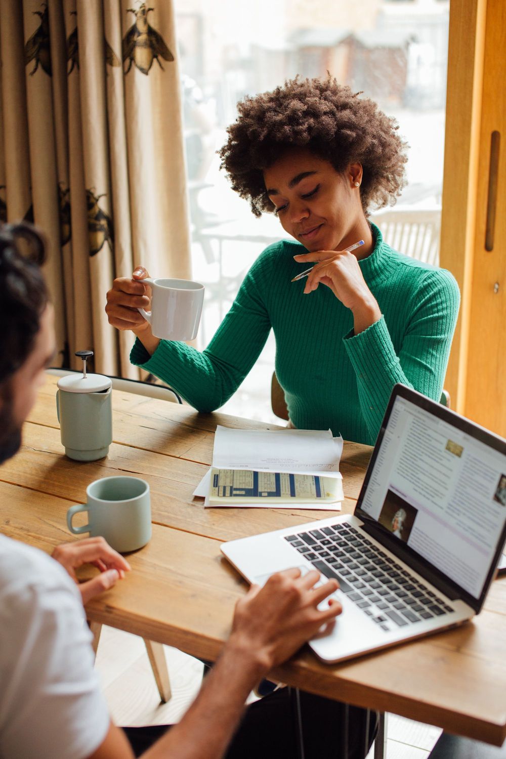 mulher negra com um café na mão olhando números, de frente para um homem branco num computador. Ambos tomando café.