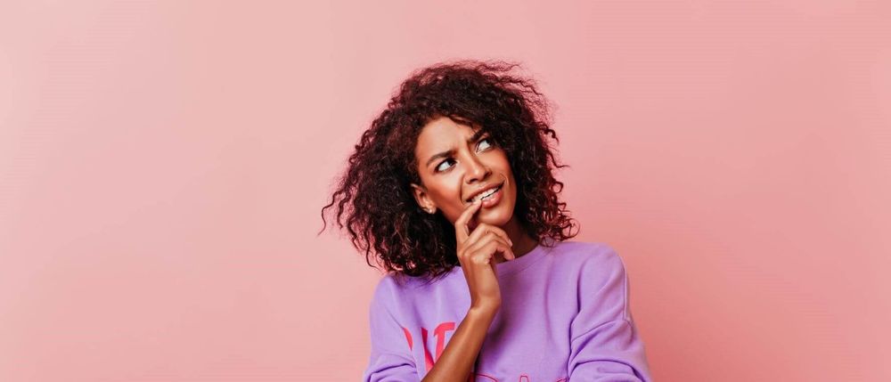 mulher negra com blusa roxa num fundo rosado, com a mão abaixo do queixo como se estivesse em dúvida ou pensando