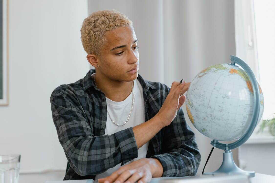 Homem negro de cabelo loiro e camisa verde listrada sentado e apoiado em mesa olhando um globo do planeta terra