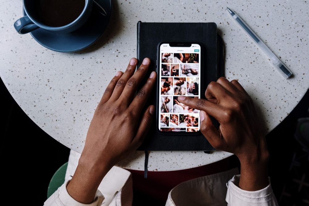 Detalhe de mãos negras tocando fotos na tela de um celular que está sobre uma mesa oval 