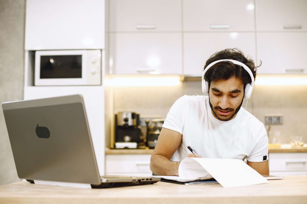 Homem de pele clara fazendo anotações em folhas brancas. Ele usa fones de ouvido e está em mesa onde há um notebook.