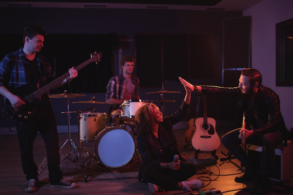 Uma banda com 4 pessoas, em uma sala. Uma mulher segurando no microfone sentada no chão, batendo na mão de um homen que toc 