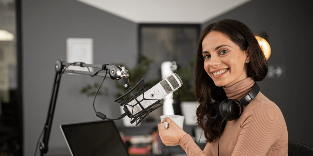 Mulher branca sentada em mesa de madeira com computador e microfone de podcast